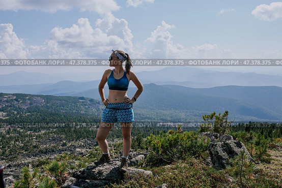 Tourist Woman in blue shorts and sports bra stands on a stone against the backdrop of summer landscape in the mountains. Стоковое фото, фотограф Екатерина Якименко / Фотобанк Лори