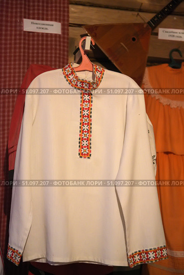 Traditional Russian folk shirt of white color, decorated with embroidery. The embroidery is done in red-black-orange tones and is located along the stand-up collar, on the chest and cuffs. The ornament is a geometric pattern with elements of crosses and diamonds, typical for Slavic motifs. The shirt hangs on a hanger against the background of the interior of a museum or ethnographic exhibition, where a balalaika and other elements of folk life are also visible. The product symbolizes the traditions, culture and originality of the Russian people. Стоковое фото, фотограф Анна Кривицкая / Фотобанк Лори