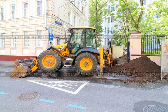 Трактор ведет ремонтные работы на улице города. Работы по прокладке коммуникаций. Экскаватор с ковшом и отвалом копает траншею (2024 год). Редакционное фото, фотограф Константин Григорьев / Фотобанк Лори