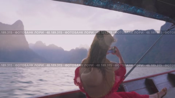 Traveling on lakes, rivers, islands and seas in Asia and Thailand. A young woman in a red dress shoots stunning views Cheow Lan Lake on her phone from a longtail boat, sailing past magnificent cliffs. Стоковое видео, видеограф Владимир Разгуляев / Фотобанк Лори