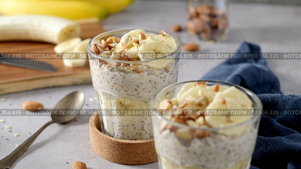Two bowls of overnight oats topped with banana slices and almonds. A spoon, cutting board, knife, and a blue napkin rest nearby. Стоковое фото, агентство Ingram Publishing / Фотобанк Лори