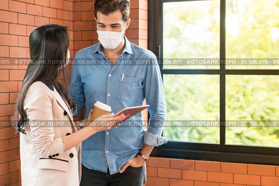 Two business person standing for brainstorm and discuss their work with digital tablet. They wear protective face mask in new normal office preventing coronavirus COVID-19 spreading. Стоковое фото, агентство Ingram Publishing / Фотобанк Лори