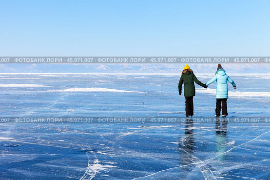 Two girls tourists skate on the blue ice of the frozen Lake Baikal near Cape Khoboy on a cold sunny February day. Beautiful winter landscape. Winter activity and outdoor recreations. Стоковое фото, фотограф Виктория Катьянова / Фотобанк Лори