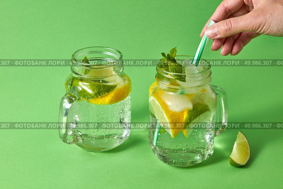 Two glass jars with cold natural cocktail homemade on green background with plastic straw with female hand. Concept of cold alcoholic or non-alcoholic summer drinks.. A woman’s hand inserts plastic straw into a glass jar with lemonade from natural ingredients - water, mint, slices of lemon, lime. Natural cold homemade drinks. Стоковое фото, агентство Ingram Publishing / Фотобанк Лори