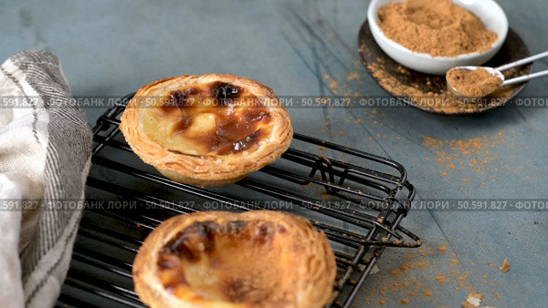 Two Portuguese custard tarts cool on a black rack next to a small bowl of cinnamon and a measuring spoon on a gray surface. Стоковое фото, агентство Ingram Publishing / Фотобанк Лори