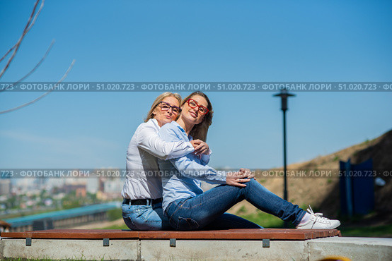 Two surprisingly similar women are sitting on a bench in an embrace and smiling. A middle-aged woman and her elderly mother. Beautiful well-groomed mother and daughter family look. Стоковое фото, фотограф Михаил Решетников / Фотобанк Лори