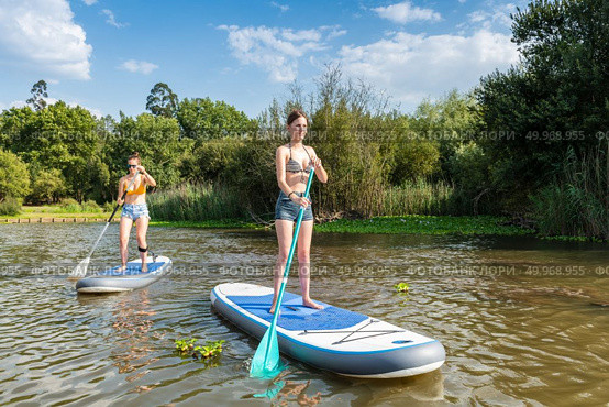 Two women stand up paddleboarding on lake. Young women doing watersport on lake. Female tourists during summer vacation. Стоковое фото, агентство Ingram Publishing / Фотобанк Лори
