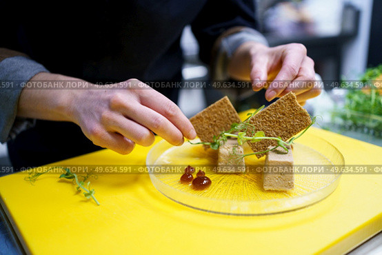 Unrecognisable chef squeezing drops of jam onto a plate of foie micuit with crunchy croutons while working in a restaurant. Crop chef plating foie micuit with crunchy croutons in restaurant. Стоковое фото, агентство Ingram Publishing / Фотобанк Лори