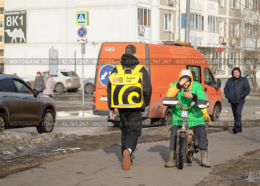 В Москве на улице встретились два курьера сервисов еды. Курьер сервиса "Яндекс.Еда" идет пешком к клиенту, а велокурьер  в форменной одежде "Сбермаркет". сидя на своем велосипеде, отдыхает со смартфоном в руках (2024 год). Редакционное фото, фотограф Наталья Николаева / Фотобанк Лори