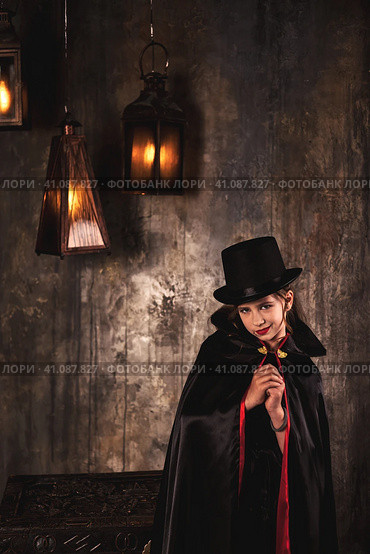 Vampire in cloak, top hat posing near chest in underground castle room, grinning at camera. Cover girl child actress in Dracula image smirking indoors. Theatre performing concept. Copy ad text space. Стоковое фото, фотограф Aleksandr Golubev / Фотобанк Лори