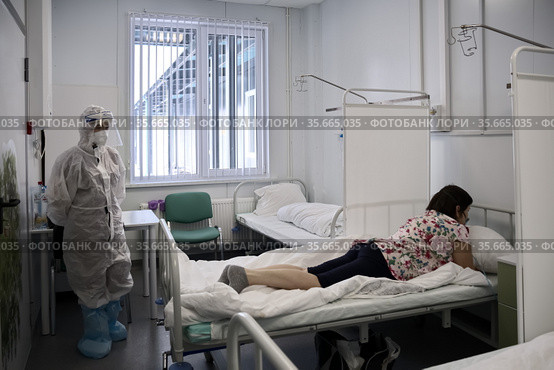 Врач в защитном костюме от COVID-19 в палате, рядом с лежащей на животе больной, в инфекционном отделении, в красной зоне медицинского центра ​помощи и спасения. Богучаны, Красноярский край, Россия (2021 год). Редакционное фото, фотограф Светлана Попова / Фотобанк Лори
