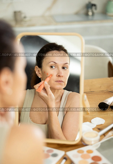 Woman applying orange concealer on cheekbones in front of vanity mirror. Стоковое фото, фотограф Яков Филимонов / Фотобанк Лори
