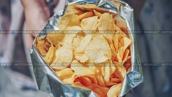 Woman grasping crinkled foil potato chip bag, displaying golden crispy snacks ready for casual outdoor munching. Стоковое фото, фотограф Mikhail Davidovich / Фотобанк Лори