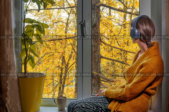 Woman in headphones and yellow knitted sweater is sitting comfortably by window with mug of cocoa. Bright autumn colors create cozy and contemplative atmosphere. Стоковое фото, фотограф Алла Чесноковва / Фотобанк Лори