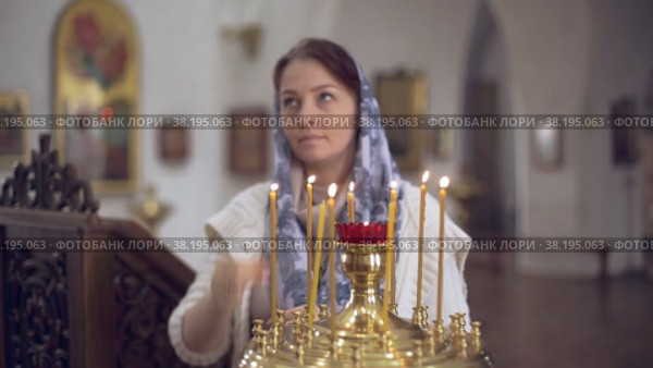 Woman in the Russian Orthodox Church with red hair and a scarf on her head lights a candle and prays in front of the icon. Стоковое видео, видеограф Арзюков Вадим Николаевич / Фотобанк Лори
