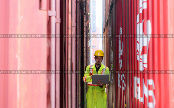 Workers in the import and export industries inspect the cargo of containers while holding a laptop and standing between them. Стоковое фото, агентство Ingram Publishing / Фотобанк Лори