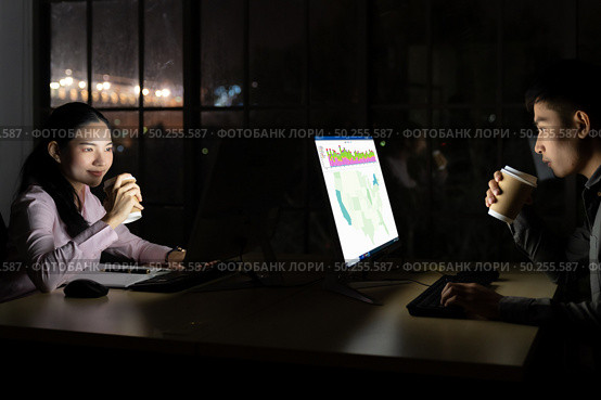 Young adult asian businessman and woman drinking hot cofee while working late at night in their office with desktop computer and laptop. Using as hard working and working late concept. Стоковое фото, агентство Ingram Publishing / Фотобанк Лори