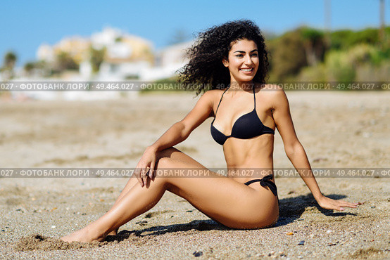 Young arabic woman with beautiful body in swimwear sitting on the beach sand. Smiling female with curly long hairstyle wearing black bikini.. Arabic woman with beautiful body in bikini lying on the beach sand. Стоковое фото, агентство Ingram Publishing / Фотобанк Лори