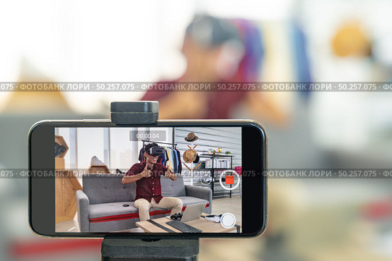 Young asian male online blogger recording live vlog video for review joystick for gaming IT product goods. Online influcencer on social media concept. Focus on mobile phone. Стоковое фото, агентство Ingram Publishing / Фотобанк Лори