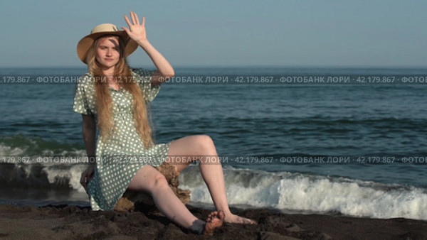 Young blonde in summer straw hat polka dots dress sits on log on shore of sea with black sand beach. Стоковое видео, видеограф А. А. Пирагис / Фотобанк Лори