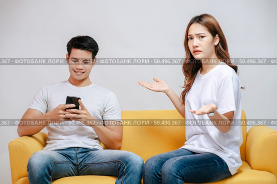 young couple arguing while handsome man using mobile phone on sofa in living room at home. husband addict On Mobile Phone Ignoring her, upset couple, family issues and relationship concept. Стоковое фото, агентство Ingram Publishing / Фотобанк Лори