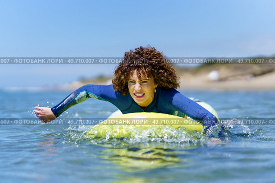 Young curly haired female surfer in wetsuit lying on SUP board and paddling with hands floating over sea against blue sky. Determined woman on paddleboard rowing with hands in ocean. Стоковое фото, агентство Ingram Publishing / Фотобанк Лори