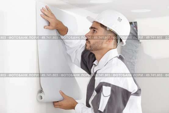 young decorator holding roll of wallpaper against a wall. Стоковое фото, агентство Ingram Publishing / Фотобанк Лори