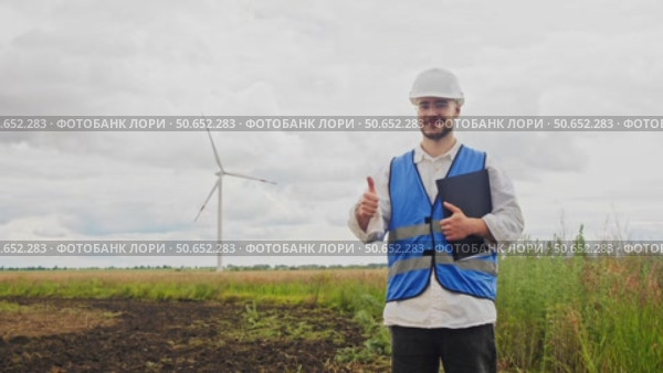 Young engineer raises his finger up and smiles at camera, thereby supporting creation of wind turbines that generate sustainable, environmentally friendly electricity. Стоковое видео, видеограф Strunov David / Фотобанк Лори