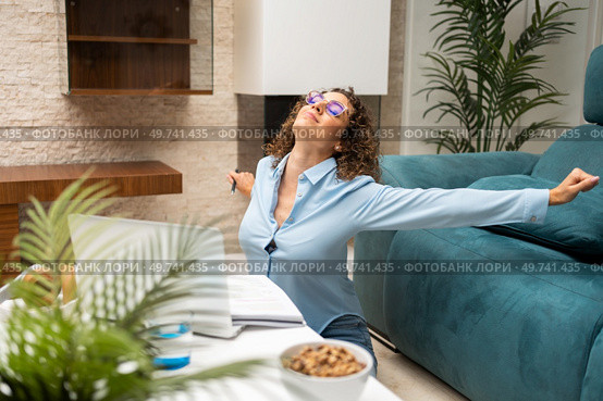 Young female freelancer in eyeglasses and smart casual stretching arms while working on laptop at table in living room at home. Happy woman stretching arms near laptop at home. Стоковое фото, агентство Ingram Publishing / Фотобанк Лори