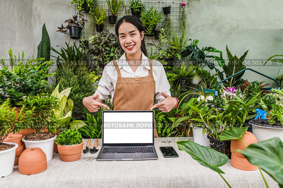Young gardener female with Mockup monitor of Laptop computer, smartphone, shovel, pitchfork, and garden equipment arrange on desk with houseplant in workplace of greenhouse. Стоковое фото, агентство Ingram Publishing / Фотобанк Лори