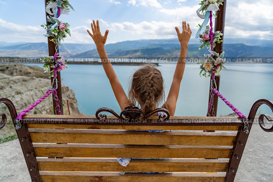 Young girl swings joyfully with her arms raised, enjoying the view of a peaceful lake and distant mountains under a clear blue sky, Смотровая площадка на Чиркейское водохранилище, Республика Дагестан, Буйнакский район, сельское поселение Чиркей. Стоковое фото, фотограф Данила Большаков / Фотобанк Лори