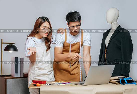 Young handsome man and pretty woman, Couple designer has conflict together while use laptop computer chatting with customer online order, mannequin and tailoring tools on desk. Стоковое фото, агентство Ingram Publishing / Фотобанк Лори