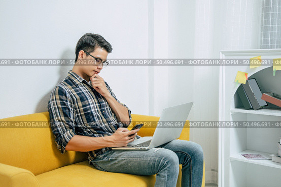 Young handsome man wearing eyeglasses with new normal, sitting on sofa and works from home on laptop computer while self-isolation and quarantine covid-19 coronavirus outbreak. Стоковое фото, агентство Ingram Publishing / Фотобанк Лори