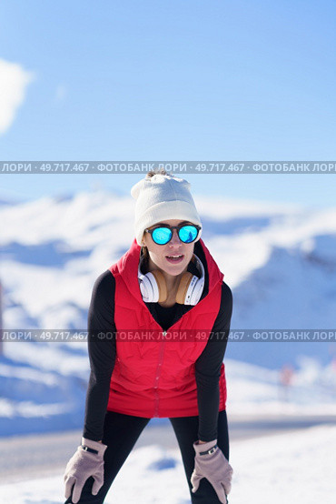 Young healthy woman in outerwear and sunglasses resting while leaning on knees against mountains in snow. Female runner in warm clothes in winter. Стоковое фото, агентство Ingram Publishing / Фотобанк Лори