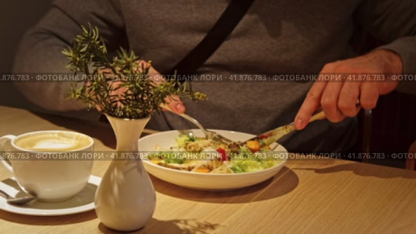 Young Man Wearing Gray Sweater Eating Fresh Vegetable Salad. Unrecognized Male Person Eating Caesar Salad in a Cafe or Restaurant and drinking Coffee while Sitting at a Wooden Table. Business Lunch. Стоковое видео, видеограф Марина Демидюк / Фотобанк Лори
