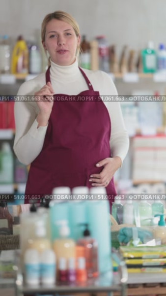 Young pregnant saleswoman wearing burgundy apron standing in personal care and cleaning supplies store aisle, smiling and gesturing warmly, welcoming customers to shopping. Стоковое видео, видеограф Яков Филимонов / Фотобанк Лори