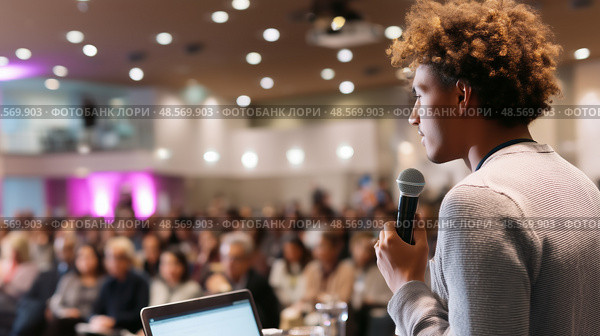 Young speaker addresses a large audience at a conference in a modern venue, showcasing public speaking skills and engagement. Стоковое AI-изображение, AI-художник Сергей Толмачёв / Фотобанк Лори