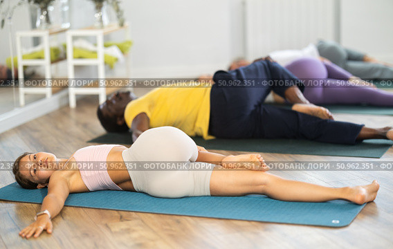 Young woman coach and students perform Supta Ardha Matsyendrasana during training in studio. Стоковое фото, фотограф Яков Филимонов / Фотобанк Лори