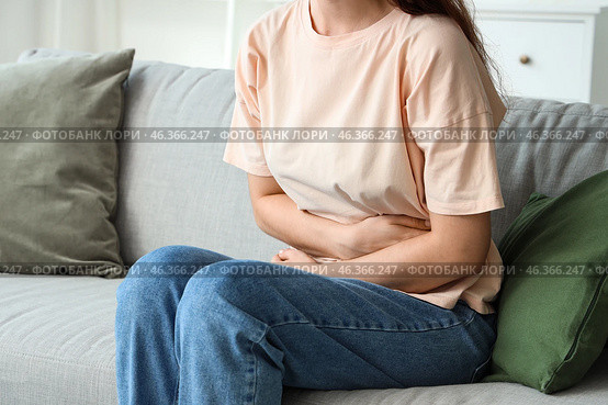 Young woman sitting on sofa and suffering from abdominal pain at home. Стоковое фото, фотограф Pixel_shot / Фотобанк Лори
