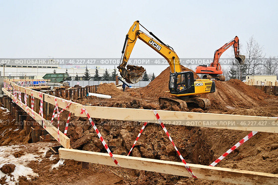 Желтый экскаватор JCB и оранжевый экскаватор работают на стройплощадке, разрабатывая грунт. В переднем плане деревянное ограждение с красно-белой лентой, траншея и строительные материалы. На заднем плане видны промышленные здания, контейнеры и строительные конструкции. (2024 год). Редакционное фото, фотограф Александр Орымбаев / Фотобанк Лори