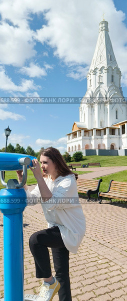 Женщина 38 лет с длинными темными волосами в белой толстовке черных штанах и белых кроссовках держит за ручки уличный синий телескоп бинокль на смотровой площадке поставив ногу на железное кольцо. за ее спиной видна белоснежная шатровая колокольня Георгия победоносца на фоне голубого неба и белых облаков. Стоковое фото, фотограф Светлана Русских / Фотобанк Лори