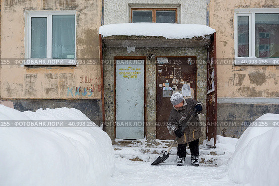 Женщина с лопатой для снега расчищает путь между сугробами. Тяжелая женская работа. Дворник убирает двор возле подъезда старого панельного дома. Магадан, Россия. Редакционное фото, фотограф Андрей Степанов / Фотобанк Лори