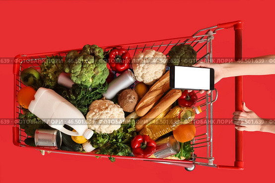 Женщина с мобильным телефоном толкает тележку, полную продуктов, на красном фоне. Стоковое фото, фотограф Pixel_shot / Фотобанк Лори